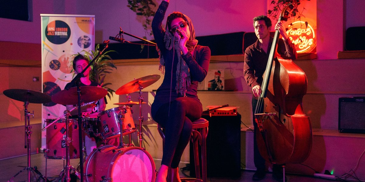 Judi Jackson wearing sunglasses singing whilst sitting on a chair with drummer and bassist in background, lit by pink light