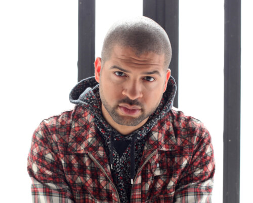 Black man in plaid shirt and jeans sits with hands meeting infront