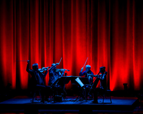 Blue lit string quartet playing wildly against a bright red curtain