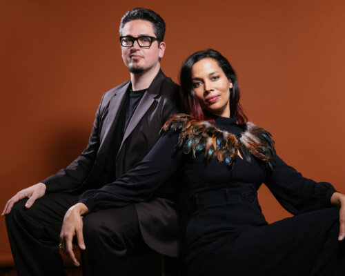Multiethnic lady in black clothes and feather shrug sits and leans on Italian man in dark suit and black glasses sitting behind