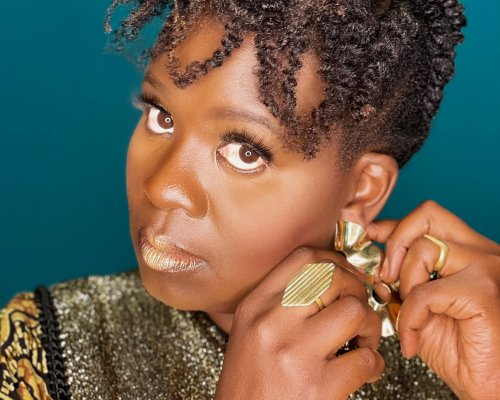 Black woman wearing gold patterns adjusts gold earring in front of teal background