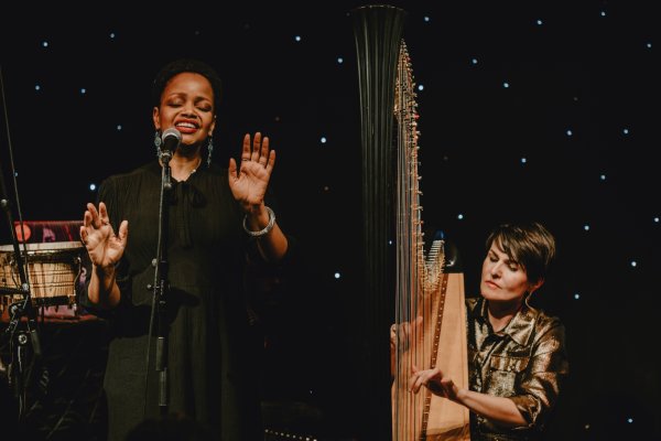 Vocalist Vimala Rowe, wearing black, passionately sings into a microphone, next to harpist Alina Bzhezhinska who wears a bronze shirt, and strums her harp with intent focus.