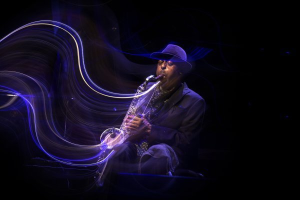 Archie Shepp sits down in a dark bowler hat and suit, playing his saxophone passionately. Streams of blue, white and purple light emulate from his instrument.