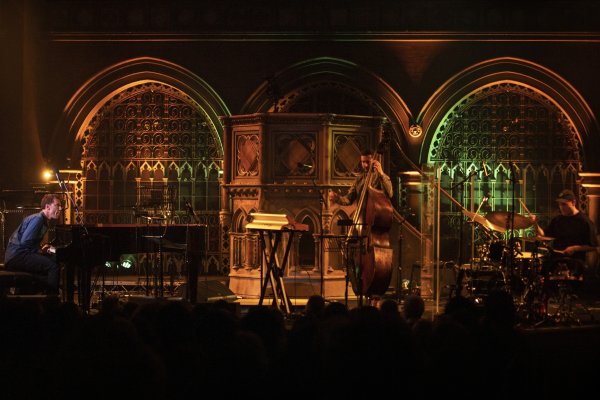 Bill Laurance plays the piano intently on the left wearing a blue shirt, joined by a bassist in the middle-ground, and a drummer on the left, wearing a baseball cap and black t-shirt. The chapel backdrop is lit up by a warm orange light.