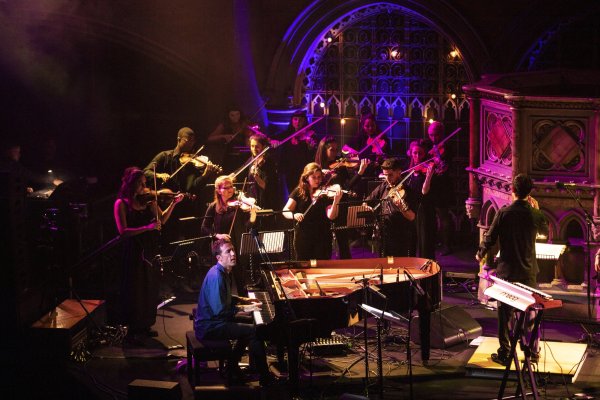 Pianist Bill Laurance, dressed in an iridescent blue shirt, plays in front of an orchestra of violin players dressed in black. They are illuminated by pink and purple lights in an old chapel setting.