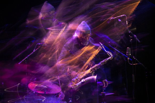 Bearded saxophonist Charles Lloyd wears a grey beanie hat and shirt, and plays centre-stage. He is immersed in a blur of orange and purple streams of light.