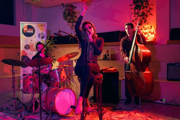 Judi Jackson wearing sunglasses singing whilst sitting on a chair with drummer and bassist in background, lit by pink light