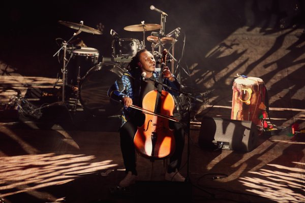 Cellist Abel Selacoe, wearing a patterned blue and black jacket and white trainers, performs in front of an empty drum kit. Patterned warm lights dapple the stage.