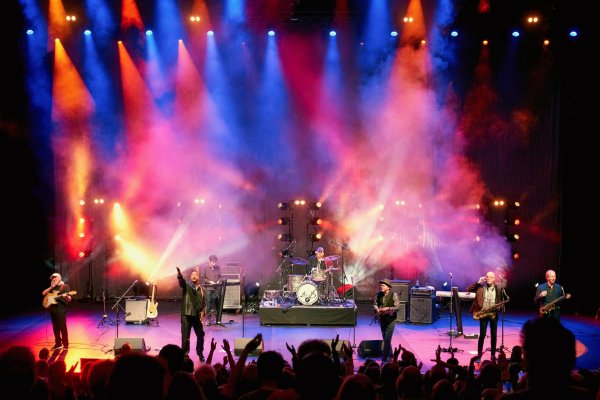Seven male band members stand on stage reaching their hand out to an audience clapping and cheering. The stage is lit up by multi-coloured flairs and smoke.