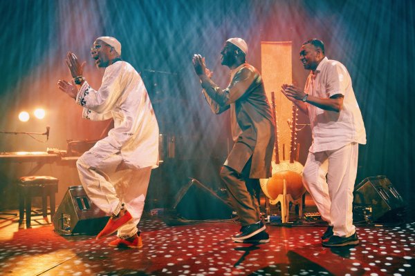 Three men dance on stage in a row, their hands poised into a clap. Two of them are wearing brown and white kaftans, and the third wears a white shirt and trousers. They are flooded with streams of white spotted lights which make a pattern on the stage.