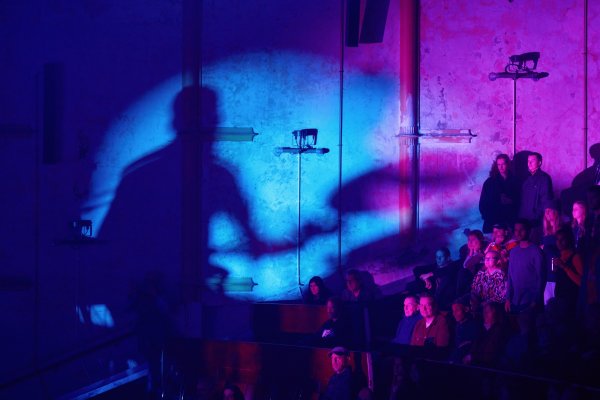 The shadow of a performing drummer projects in a blue circle onto the wall of the theatre. An audience sits beneath it.