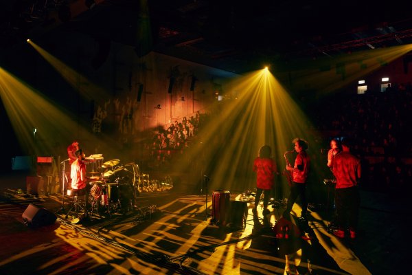 From behind the stage, Sarathy Korwar sits at a drumkit, to the left of a band of musicians. They are flooded in warm orange and yellow lights.