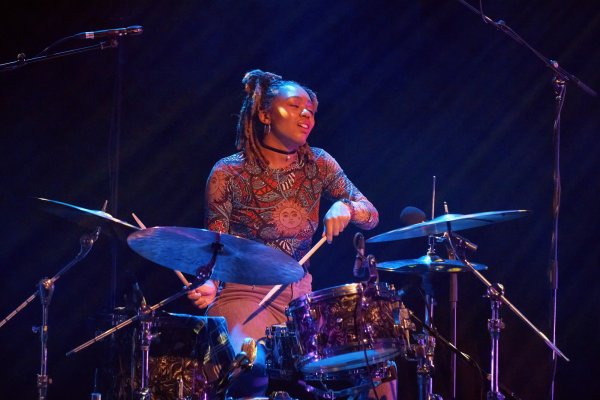 A close-up of drummer Romarna Campbell who plays her kit centre-stage, wearing a patterned top and brown trousers.