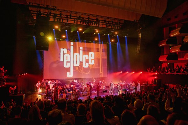 An audience gives a standing ovation to a huge band of musicians on a stage. The word "rejoice" is written on the screen behind them and red and blue lights flood the stage and audience.