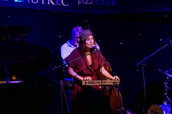 A woman wearing a purple hat and crimson dress plays the synth in front of a star-studded backdrop.