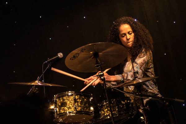 A close-up of drummer Jas Kayser who plays her kit with focus in front of a star-studded backdrop.