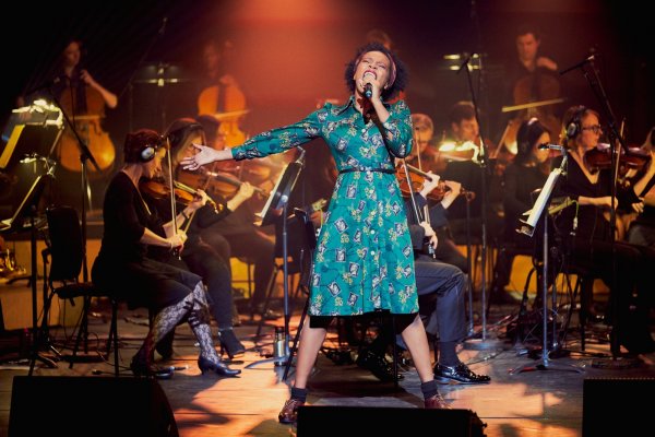 Cherise singing with eyes closed, in a star-shaped posture, wearing green patterned dress, in front of orchestra