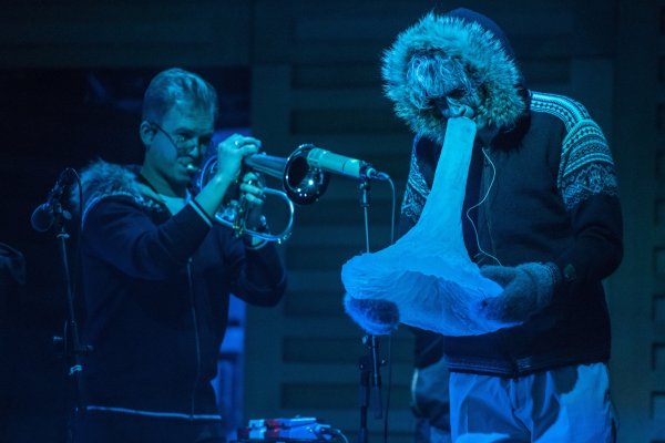 Two musicians wear furry jumpers, illuminated by blue lights. One on the left plays the trumpet, whilst the other blows into a shape of ice.