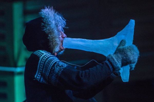 A close-up of Terje Isungset wearing a furry hood and gloves blowing into cone-like shape of ice.