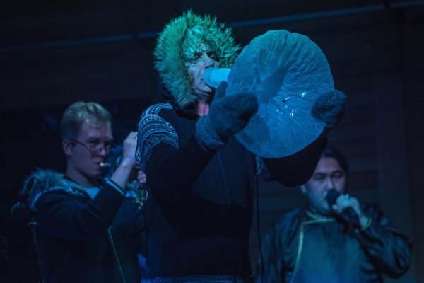 A front close-up of Terje Isungset wearing a furry hood and gloves blowing into cone-like shape of ice in front of a trumpeter and singer.