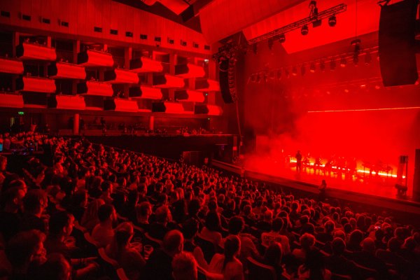 A seated audience watch onto a stage flooded with red smoke and light.