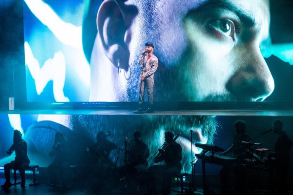 Performer Woodkid sings into a microphone wearing a grey boiler suit in front of an enlarged projection of his face. He stands on a platform above a row of the silhouettes of musicians.