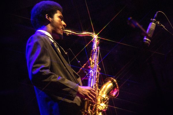 A close-up from beneath of saxophonist Xhosa Cole wearing a dark suit and playing his instrument which sparkles in the light.